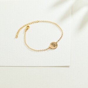 May include: A delicate gold-colored bracelet with a floral design on a circular charm. The adjustable chain features a lobster clasp closure. The bracelet is displayed on a white surface, showcasing its elegant design.