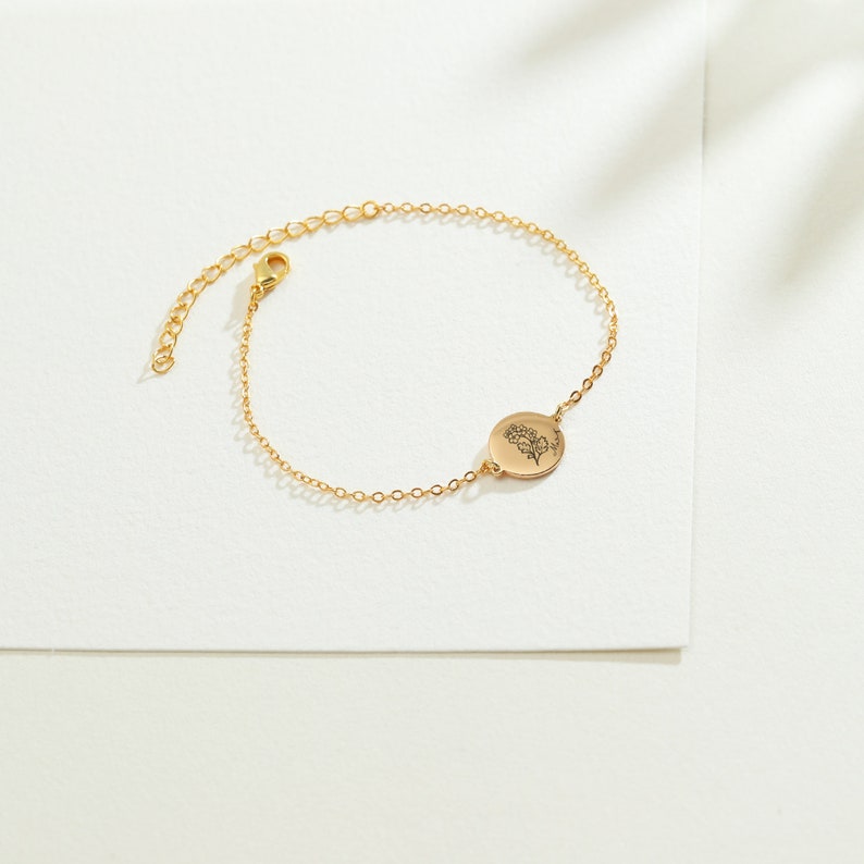 May include: A delicate gold-colored bracelet with a floral design on a circular charm. The adjustable chain features a lobster clasp closure. The bracelet is displayed on a white surface, showcasing its elegant design.
