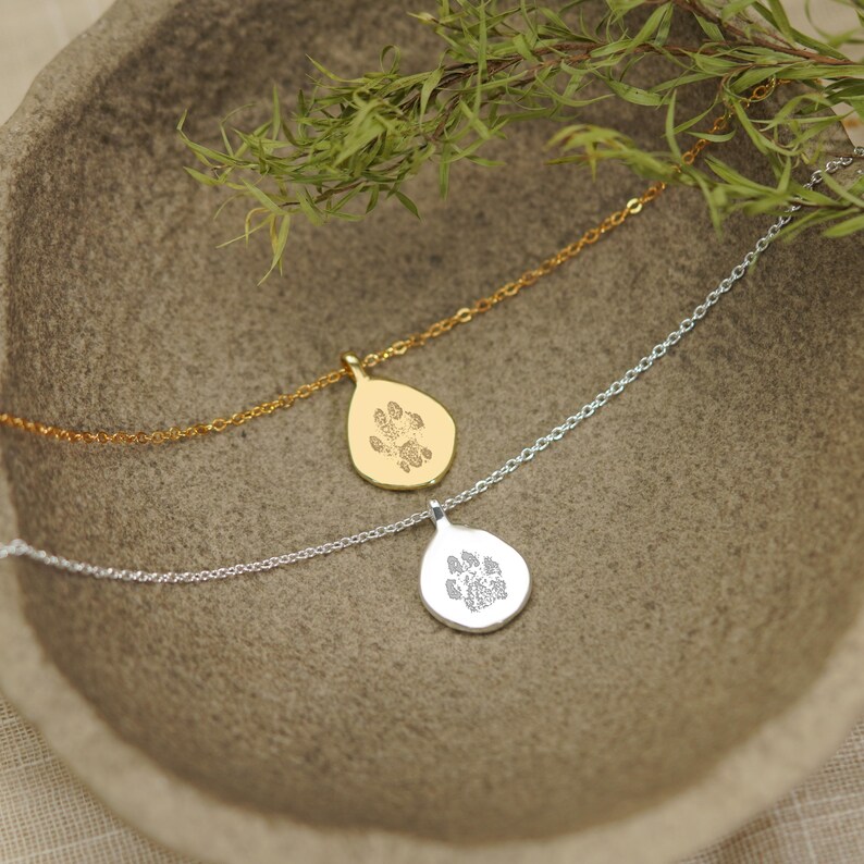 Puede incluir: Dos collares con colgantes en forma de l&aacute;grima, uno en oro y otro en plata. Cada colgante presenta un dise&ntilde;o grabado de una huella de animal. Los colgantes est&aacute;n unidos a finas cadenas de metal a juego, con un dise&ntilde;o sencillo y limpio.