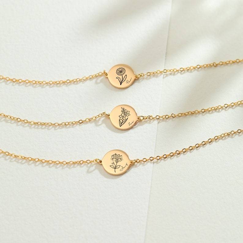 May include: Gold-colored bracelets with delicate chain links. Each bracelet features a circular charm with a floral design and a handwritten name. The charms are engraved with different flowers and names, suggesting a personalized jewelry item.