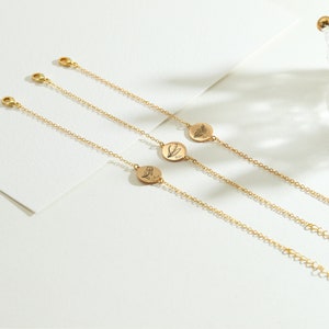 May include: Three delicate gold-colored bracelets with small, round pendants featuring floral designs. Each bracelet has a fine chain and a clasp. The pendants have engraved botanical illustrations. The bracelets are displayed on a white surface.