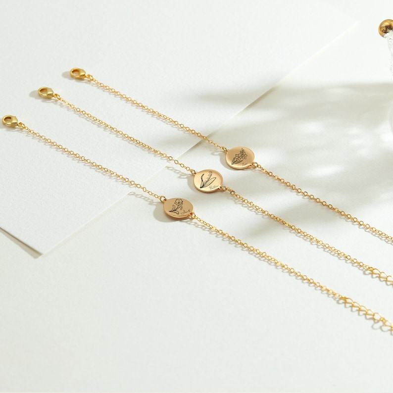 May include: Three delicate gold-colored bracelets with small, round pendants featuring floral designs. Each bracelet has a fine chain and a clasp. The pendants have engraved botanical illustrations. The bracelets are displayed on a white surface.