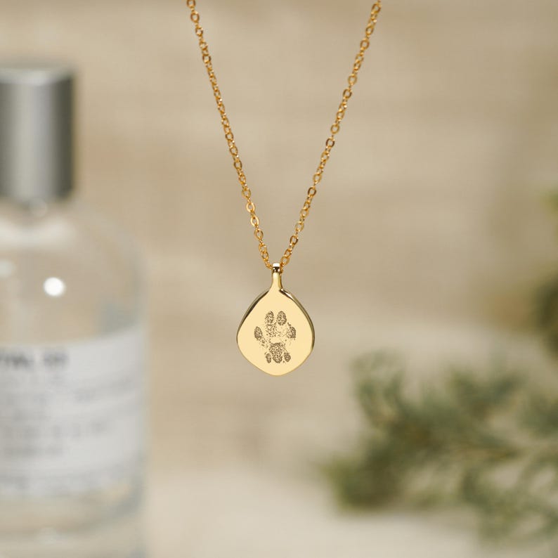 Puede incluir: Un collar de tono dorado con una cadena delicada y un colgante en forma de l&aacute;grima. El colgante muestra un grabado detallado y oscuro de una huella de pata de animal centrado en su superficie met&aacute;lica lisa y pulida.