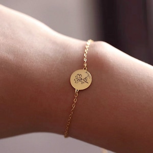 May include: A gold-colored bracelet featuring a delicate chain and a circular pendant. The pendant is engraved with a floral design and script. The bracelet is shown on a person's wrist, highlighting the jewelry's design and details.