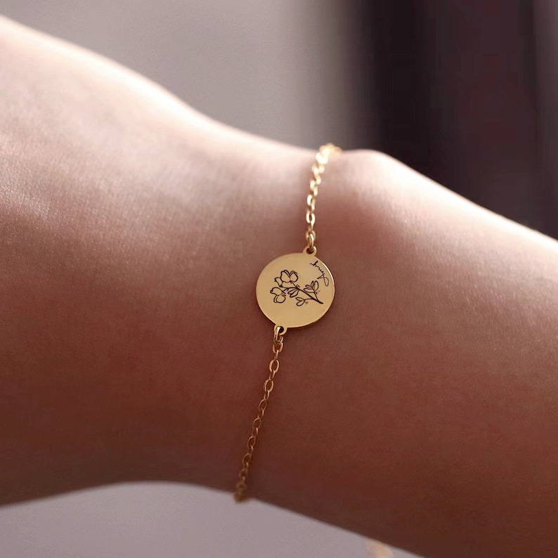 May include: A gold-colored bracelet featuring a delicate chain and a circular pendant. The pendant is engraved with a floral design and script. The bracelet is shown on a person's wrist, highlighting the jewelry's design and details.