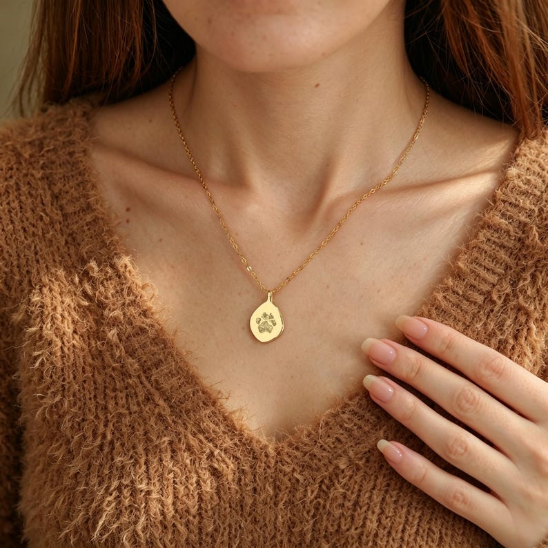 Puede incluir: Un collar de tono dorado con una cadena delicada y un peque&ntilde;o colgante de forma org&aacute;nica e irregular. El colgante presenta un grabado detallado de una huella de pata centrado en su superficie lisa y pulida, sobre una tela marr&oacute;n texturizada.