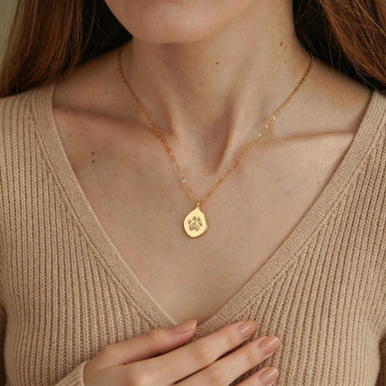 Puede incluir: Un collar de tono dorado que presenta una cadena delicada y un peque&ntilde;o colgante con forma de gota irregular. El colgante est&aacute; grabado con un dise&ntilde;o de huella de pata de animal centrado en su superficie lisa y pulida, colgado de una cadena fina.