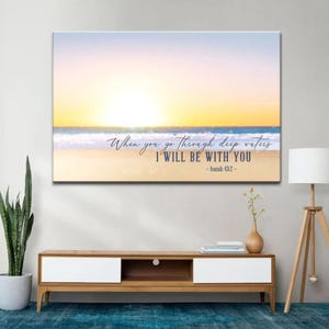 May include: A canvas print depicting a beach scene at sunset. The artwork features a vibrant sky with shades of yellow and orange, a blue ocean, and a sandy beach. Text reads "When you go through deep waters I WILL BE WITH YOU - Isaiah 43:2".