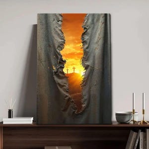 May include: A canvas print depicting three crosses on a hill at sunset, framed by a torn, textured fabric. The sky is a vibrant orange and yellow, creating a dramatic and spiritual scene. The artwork is displayed on a dark wooden shelf.