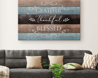May include: A wooden wall art piece with the words "Grateful, Thankful, Blessed" in white script. The background features horizontal planks in shades of brown, blue, and black, creating a rustic aesthetic. Decorative swirls and leaves accent the text.