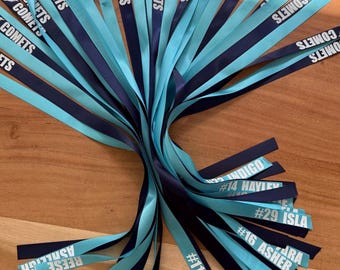 May include: A collection of satin ribbons in alternating light blue and dark blue colors. The ribbons feature the printed text COMETS, along with various names and numbers such as #11 RUBY, #16 ASHER, #29 ISLA, #14 HAYLEY, and INDIGO.