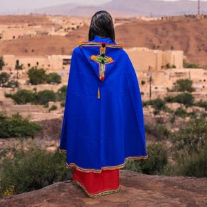 May include: A person wearing a vibrant blue cape with a decorative cross design on the back. The cape has a red trim and a gold tassel. The person is standing in front of a scenic landscape with buildings and trees.