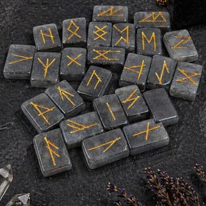 Puede incluir: Un conjunto de piedras rúnicas grises con símbolos dorados, dispuestas sobre una superficie oscura y texturizada. La imagen incluye una vela encendida, un libro encuadernado en cuero, una bolsa de terciopelo negro y cristales, sugiriendo un tema místico.