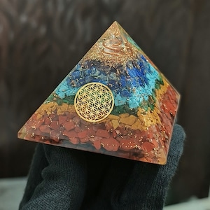 Peut inclure: Une pyramide de cristal à sept chakras avec un symbole doré de la fleur de vie au centre. La pyramide est faite de cristaux rouges, orange, jaunes, verts, bleus, indigo et violets.