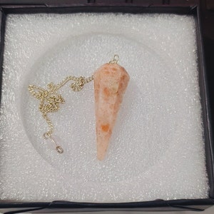 Op de afbeelding: Een gouden ketting met een zonnesteenpendulum. Het pendulum is licht oranje van kleur met donkerdere oranje vlekken. Het bevindt zich in een witte doos met een zwart deksel.
