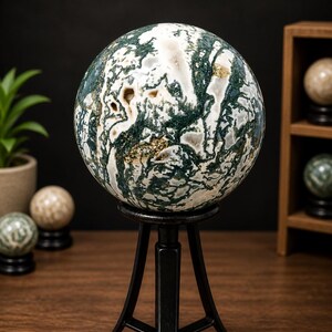 May include: A large, polished moss agate sphere, displaying intricate patterns of white, green, and brown, is displayed on a black metal tripod stand. Other spheres are visible in the background, suggesting a collection of decorative stones.