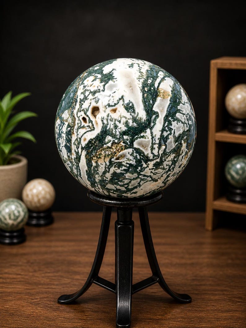 May include: A large, polished moss agate sphere, displaying intricate patterns of white, green, and brown, is displayed on a black metal tripod stand. Other spheres are visible in the background, suggesting a collection of decorative stones.