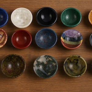 Puede incluir: Una vista cenital muestra una colección de cuencos pequeños y redondos en varios colores y patrones. Los cuencos están dispuestos sobre una superficie de madera, mostrando una diversa gama de tonos, incluyendo azul, blanco, rojo y verde. Los cuencos parecen ser de piedra o cerámica.