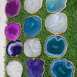May include: A collection of 15 agate coasters with gold trim. The coasters are in various shades of purple, blue, and white. They are arranged on a green grassy surface.