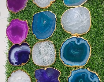 May include: A collection of 15 agate coasters with gold trim. The coasters are in various shades of purple, blue, and white. They are arranged on a green grassy surface.