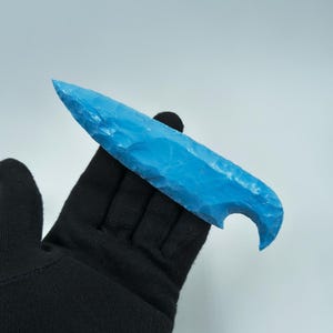 Könnte beinhalten: Ein hellblaues, handgehaltenes, flintartiges Objekt mit einer gebogenen Kerbe an der Basis. Das Objekt wird von einer schwarzen, behandschuhten Hand vor einem weißen Hintergrund gehalten. Das Objekt hat eine scharfe, spitze Spitze.