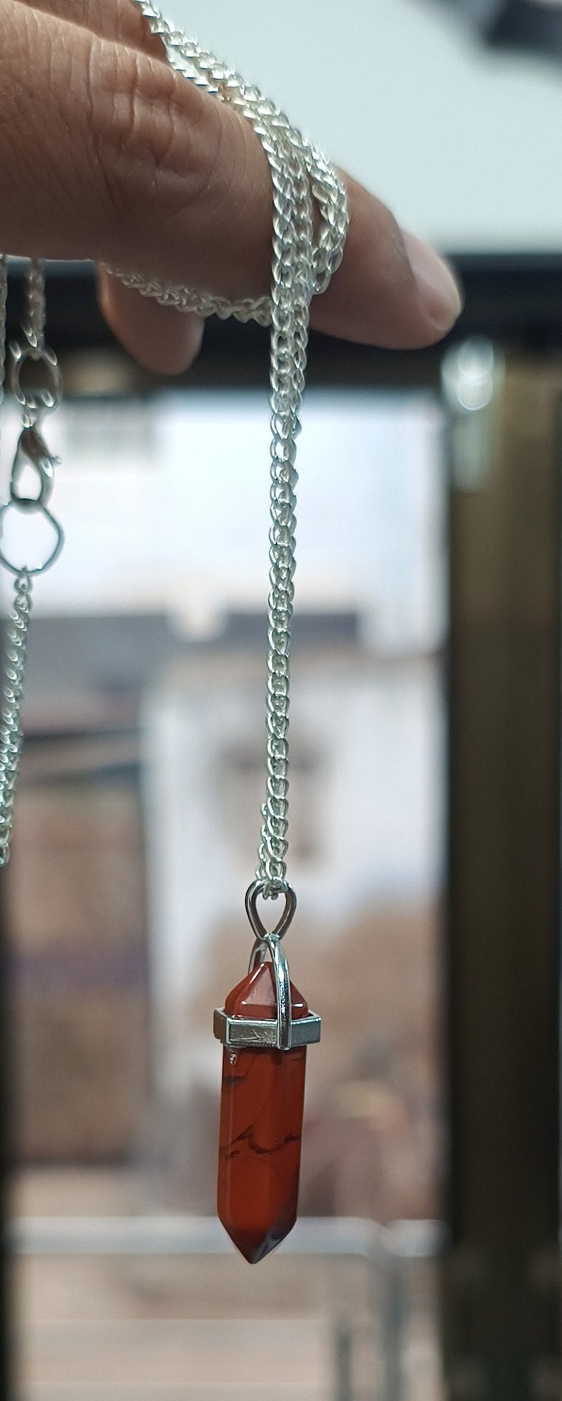 May include: A silver chain necklace with a red crystal point pendant. The pendant is a long, thin, pointed crystal with a silver setting.