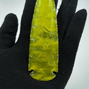 May include: A translucent, yellow-green arrowhead with a textured surface. The arrowhead is held by a gloved hand against a white background. The arrowhead has a pointed tip and a notched base.