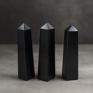 Op de afbeelding: Drie zwarte obsidiaan obelisken met een gepolijste afwerking. Elke obelisk heeft een rechthoekige basis en een puntige, piramidale top. De objecten worden getoond op een grijs oppervlak tegen een donkere achtergrond.