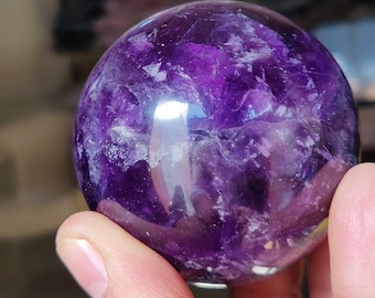 Peut inclure: Une grande sphère d'améthyste violette avec une surface lisse et polie. La sphère est translucide et présente un motif tourbillonnant de blanc et de violet.