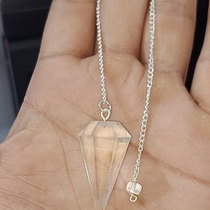 May include: A clear quartz crystal pendulum with a silver chain. The pendulum is hanging from a person's hand.