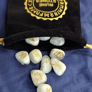 May include: A set of white rune stones with gold lettering, scattered on a blue fabric background. The stones are in a black velvet bag with a gold label that reads "Rune Stones".