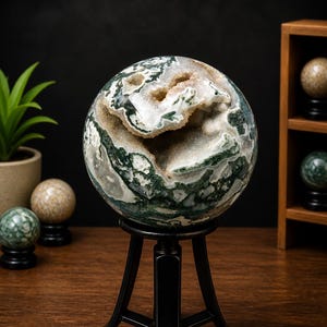 May include: A large, spherical moss agate geode with green, white, and tan patterns, displayed on a black metal stand. The geode has a hollow center with visible crystal formations. Other decorative spheres are in the background.
