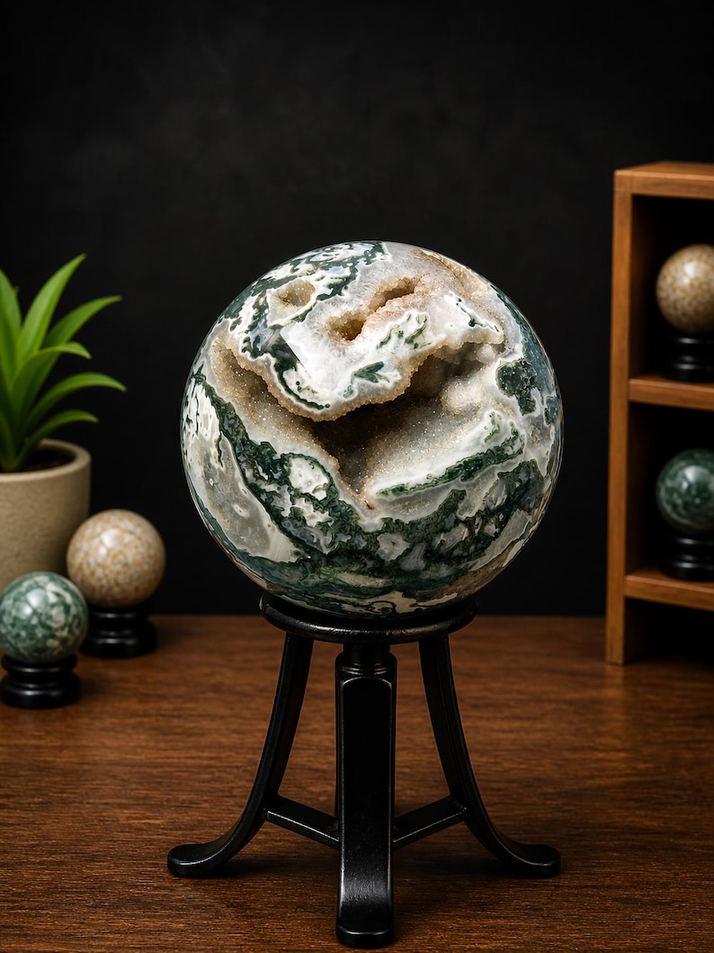 May include: A large, spherical moss agate geode with green, white, and tan patterns, displayed on a black metal stand. The geode has a hollow center with visible crystal formations. Other decorative spheres are in the background.