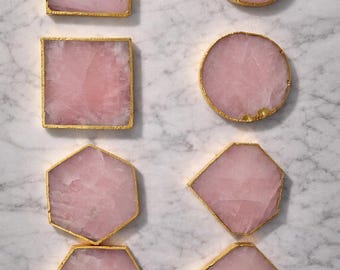 May include: Pink stone coasters in various shapes: square, round, hexagon, and diamond. Each coaster has a gold-colored edge. The coasters are arranged on a white and gray marble surface. A decorative home item.