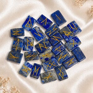 May include: A collection of rectangular, lapis lazuli-colored rune stones with gold-colored etched symbols. The stones are arranged on a cream-colored, silky surface, with dried floral accents.