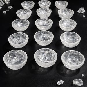 May include: A collection of clear quartz bowls arranged on a black surface. The bowls are small, round, and translucent, with visible internal veining. Several quartz crystals are scattered around the bowls, and a few green leaves are visible in the background.