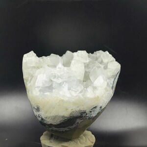 May include: A geode crystal cluster with a light blue interior and a rough, textured exterior. The geode is mounted on a carved, light brown wooden base. The crystal formation is the focal point, creating a natural aesthetic.