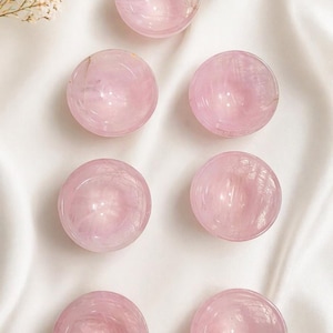 May include: Seven polished rose quartz spheres, each with a soft pink hue and natural veining. The spheres are arranged on a white, silky fabric, creating a serene and elegant display. The spheres are a popular choice for crystal healing and meditation.