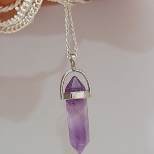 Puede incluir: Un collar de cadena de plata con un colgante de cristal de amatista púrpura. El cristal está en un engaste de plata y tiene una punta puntiaguda.