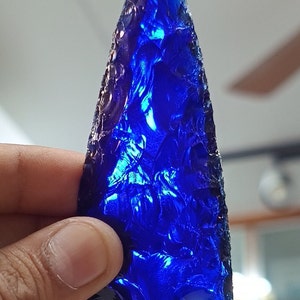 May include: A deep blue, hand-crafted arrowhead made of glass. The arrowhead has a sharp point and a textured surface.