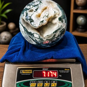 May include: A large, spherical moss agate gemstone, displaying green, white, and tan patterns. The agate rests on a blue cloth atop a digital scale, which reads 7.174 kg. The scale is silver with black and green accents. The gemstone is a decorative item.
