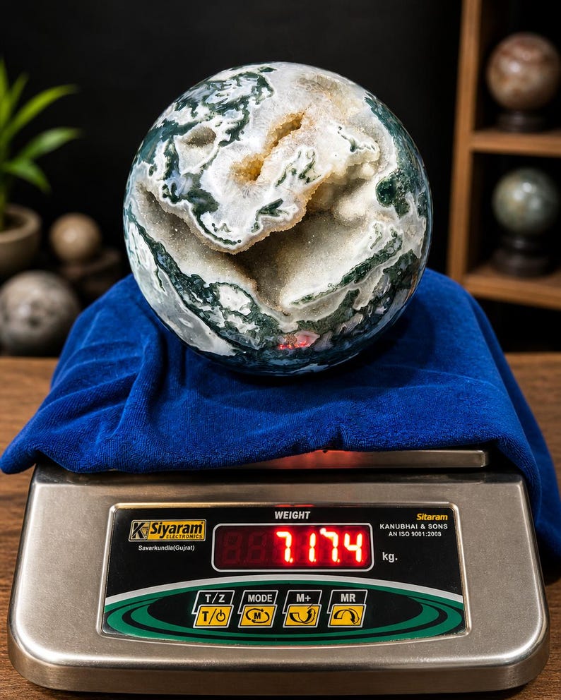 May include: A large, spherical moss agate gemstone, displaying green, white, and tan patterns. The agate rests on a blue cloth atop a digital scale, which reads 7.174 kg. The scale is silver with black and green accents. The gemstone is a decorative item.