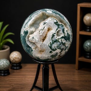 May include: A large, spherical decorative stone with green and white patterns, displayed on a black metal stand. The stone has a geode-like interior. Other decorative spheres are visible in the background.