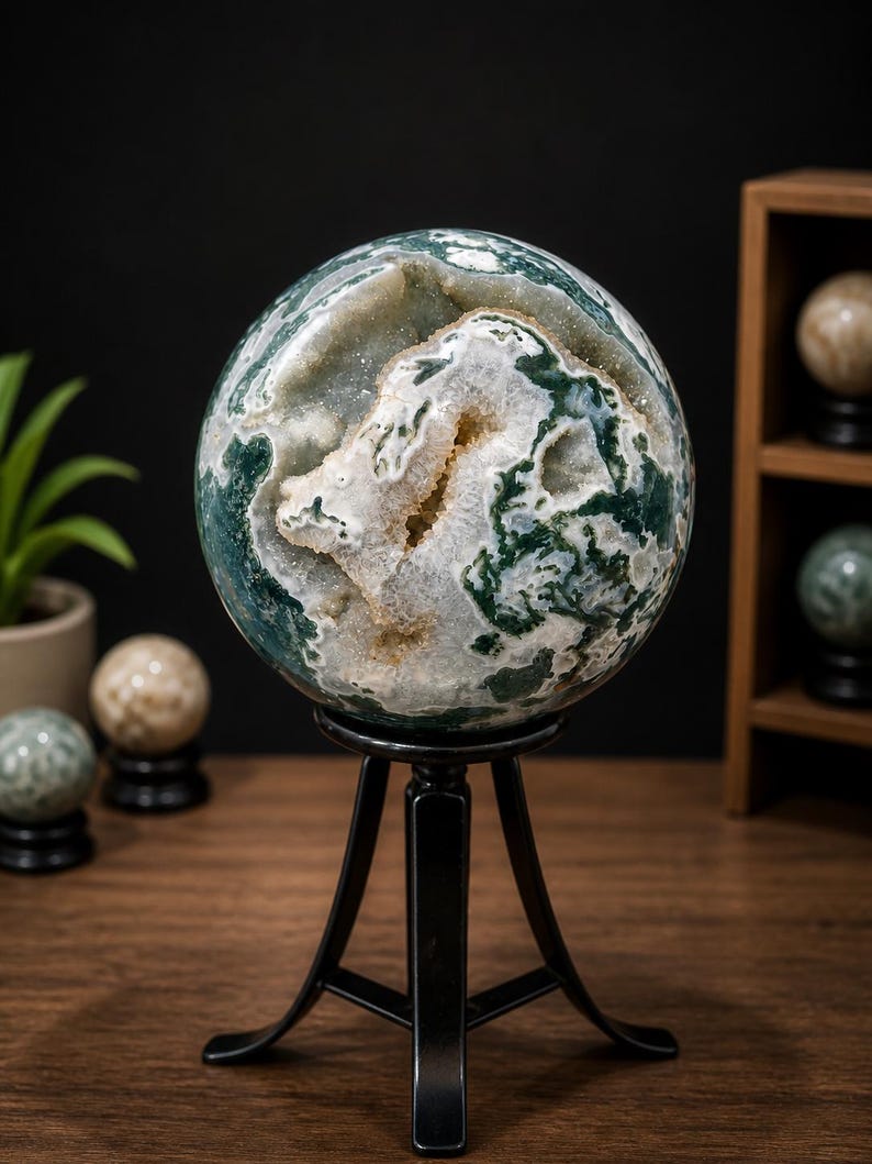 May include: A large, spherical decorative stone with green and white patterns, displayed on a black metal stand. The stone has a geode-like interior. Other decorative spheres are visible in the background.