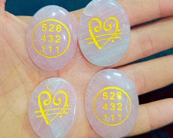 Puede incluir: Cuatro piedras ovaladas de cuarzo rosa. Dos piedras presentan un diseño de corazón dorado, y las otras dos tienen los números 528, 432 y 111 en un círculo dorado. Las piedras son de color rosa claro.