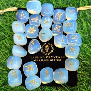 May include: A set of 18 white opal rune stones with gold runes on a black velvet pouch. The pouch has the text "Tashah Crystals New Age Healing Stone".