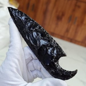 Puede incluir: Una punta de flecha de obsidiana negra con un diseño único en forma de media luna. La punta de flecha es lisa y pulida, con un acabado brillante.