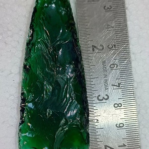 May include: A green glass arrowhead, measuring approximately 6.5 cm in length, is shown next to a ruler.