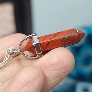 May include: A red jasper crystal pendant with silver accents on a silver chain. The pendant is shaped like a point and has a red and brown marbled pattern.