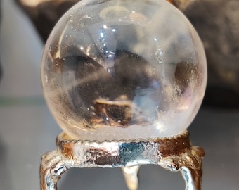 May include: A clear crystal ball on a silver stand with three legs. The ball is smooth and reflective.